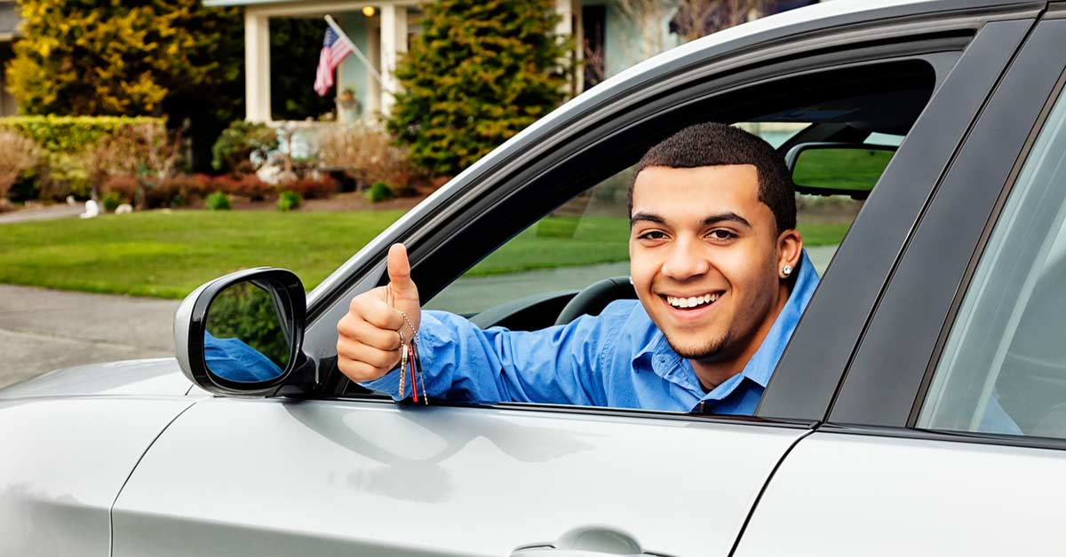 Image shows a teen driver holding a thumbs up while inside of a car smiling.